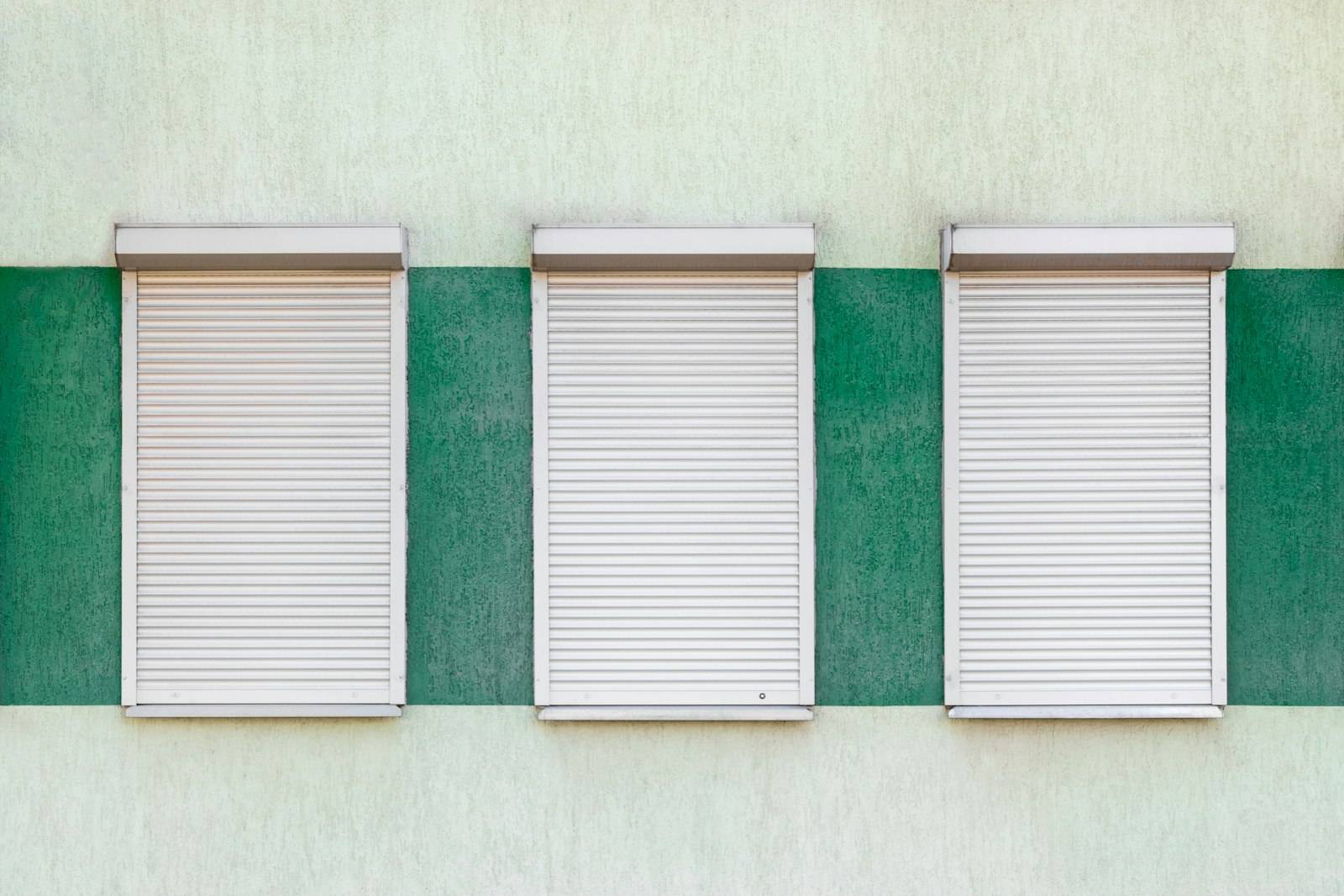 Trois fenêtres aux volets roulants blancs baissés sur une façade blanche et verte